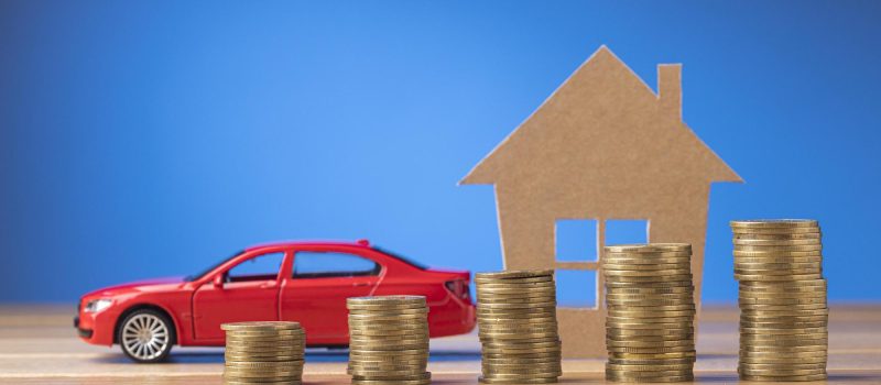 Rotes Auto neben einem Haus. Vor dem Auto sind Geldmünzen. Dachboxen. Mieten. Kaufen. Sparen. Up & Drive.